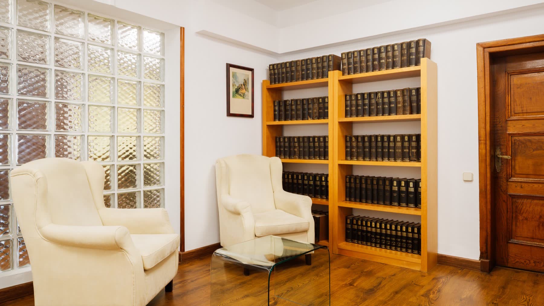 Sala con dos butacas enfrentadas, mesa baja de cristal y estantería de libros junto a una ventana de pavés