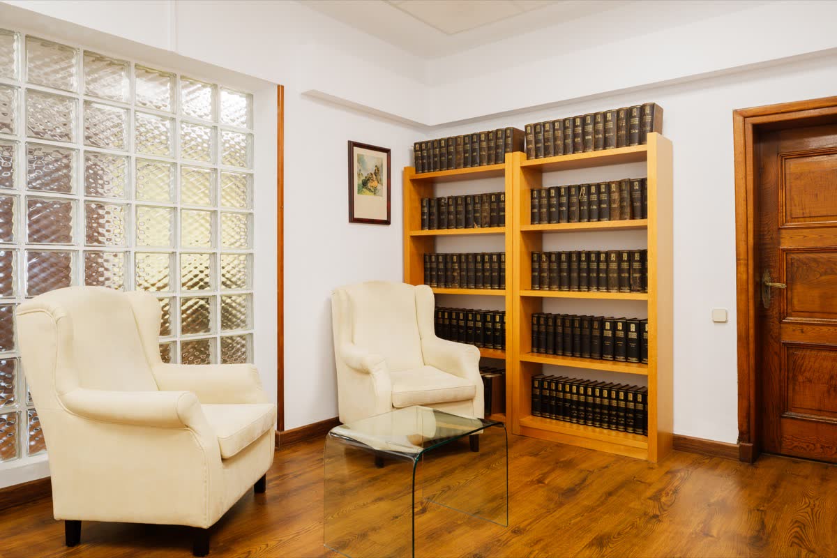 Sala con dos butacas enfrentadas, mesa baja de cristal y estantería de libros junto a una ventana de pavés