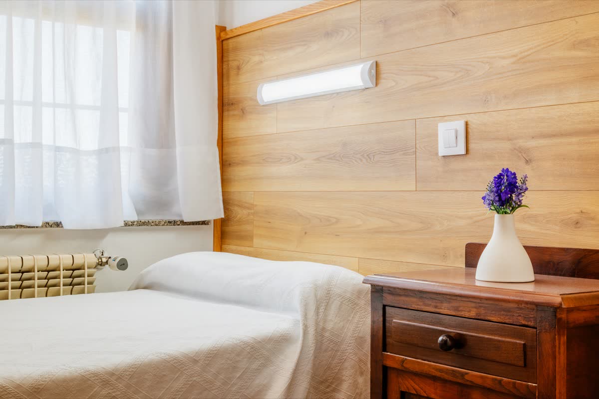 Habitación individual con cabecero de madera, mesilla con flores y ventana con visillos